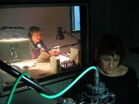 A woman sits in front of a microphone in a recording booth, and through it's window we see another woman sitting at a desk with a computer and microphone