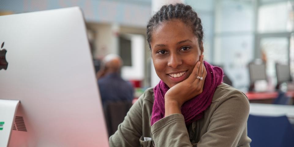 Photo of a woman smiling at the camera while sitting in front of a desktop computer.