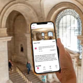 Hand holding a phone displaying the audio tour with the white marble Astor Hall in the background.