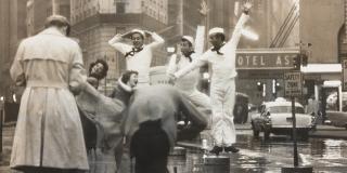 Two photographers are shown taking photos of a group of three Navy men in their white uniforms and two women. The women are dancing and the three Navy men are making faces and jumping into the air, arms stretched upward. 