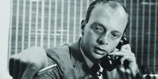 Black and white photo of man sitting at desk speaking on a rotary phone; one hand is reaching across desk and pointing a pen at an open notebook.