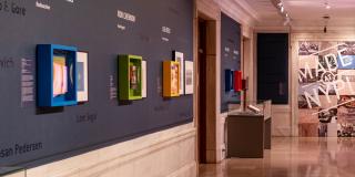 A long hall is pictured. Small, colored display cases holding books and images line the wall on the left side. A pair of display screens and glass display box on a table are shown on the right.  