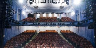 The seating of a two tier theatre is pictured from the perspective of the stage. Two large spotlight shine down on the fading red velvety seats 