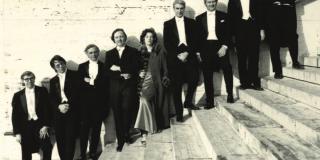 A line of musicians stand in a descending line on concrete steps against a concrete wall. 