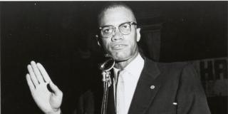 Malcolm X is shown standing at a podium. A sign attached to the podium's front says 'Marcus Garvey Day, Fri, Aug 1, 1958'