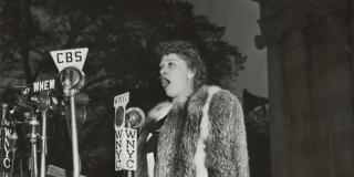 A black and white photo of a woman outside singing into several press microphones.