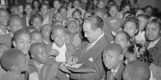 Black and white photo of Langston Hughes in a suit signing an autograph surrounded by a sea of black children