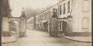 A vintage photograph in sepia tone of an urban setting, featuring a street flanked by two arched gateways and a row of townhouses