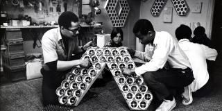 Black and white photo of two men holding each side of a large object with a woman in the background, inside a artist studio