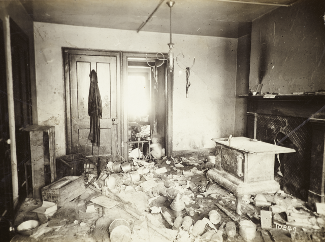 Historic black-and-white photo of a chaotic room inside of a New York City tenement with debris littered all over the floor, a wood-burning stove, an overturned chair, and a dusty jacket hung on a door; small naked child squats in a doorway in one of the few spaces where the floor is visible.