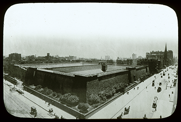 Black-and-white lantern slide of the Croton Reservoir with hedges on around the sides of the reservoir and a view of Fifth Avenue filled with carriages and pedestrians.