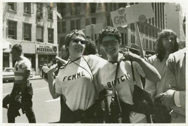 Black and white photograph of two women in march, wearing t-shirts: one says "butch," one says "femme"