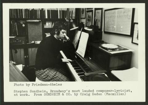 A black and white photo of Stephen Sondheim sitting at a piano in an office space