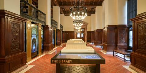 Interior of the Visitor Center at the Stephen A. Schwarzman Building, featuring tables of digital displays and touchable/tactile objects, chandeliers hanging from the ceiling, and panels of carved wood lining the walls.