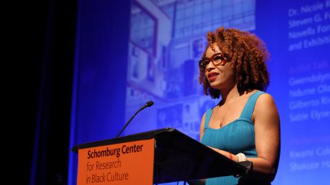 Black woman in a turquoise dress standing at a podium with an orange sign and blue background image.