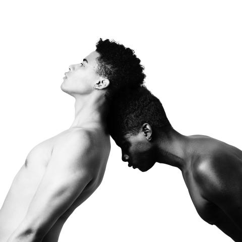 Black and White photo of two young men, one of light complexion falling back onto the head of a darker complexion young man leaning forward.