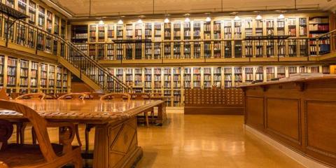 Photo of a library reading room featuring wooden desks and wall stacks of books divided into two levels