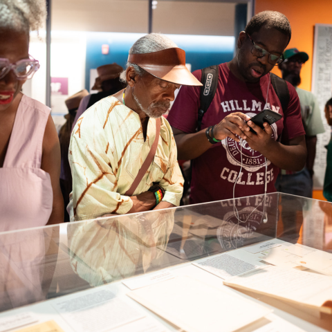 Color photo of two men and a woman viewing a display case of archival materials. One man has a cellphone in his hand with headphones.