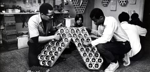 Five people kneel on the floor, two of them are in the foreground tinkering with a v-shaped contraption.